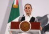 Mexico's President Claudia Sheinbaum standing at the presidential podium with two small microphones before her. She's wearing a black and gray blazer and a white turtleneck underneath.