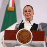 Mexico's President Claudia Sheinbaum standing at the presidential podium with two small microphones before her. She's wearing a black and gray blazer and a white turtleneck underneath.