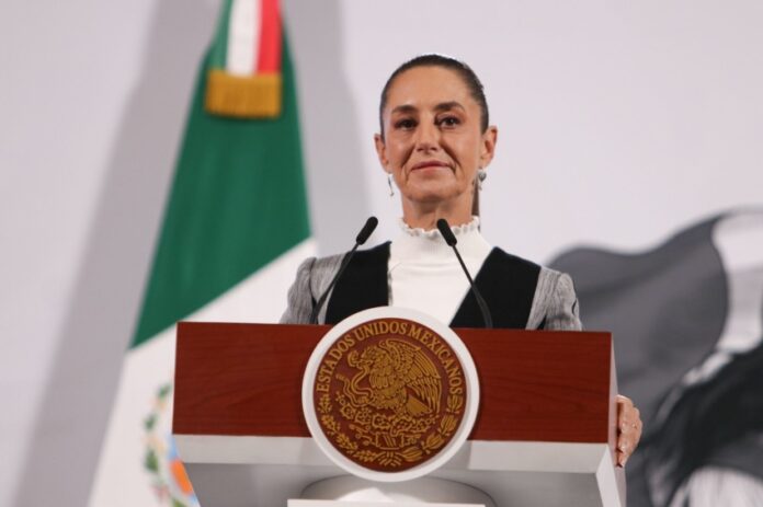 Mexico's President Claudia Sheinbaum standing at the presidential podium with two small microphones before her. She's wearing a black and gray blazer and a white turtleneck underneath.