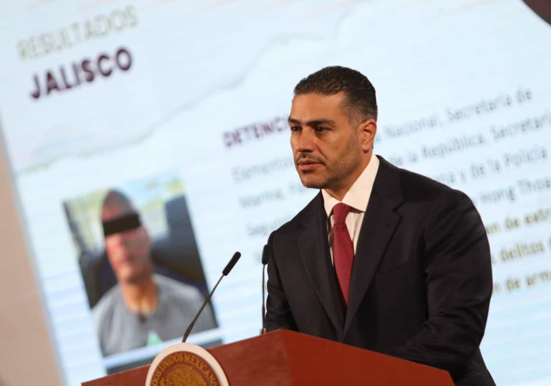 Omar Garcia Harfuch, a man in his thirties with close cropped hair, wearing a black suit and red tie stands at the presidential podium at Claudia Sheinbaum's daily press conference, giving a security report.