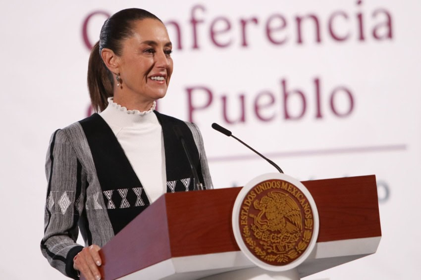 Mexico's President Claudia Sheinbaum standing at the presidential podium in the National Palace press briefing room, speaking with reporters. She is smiling.