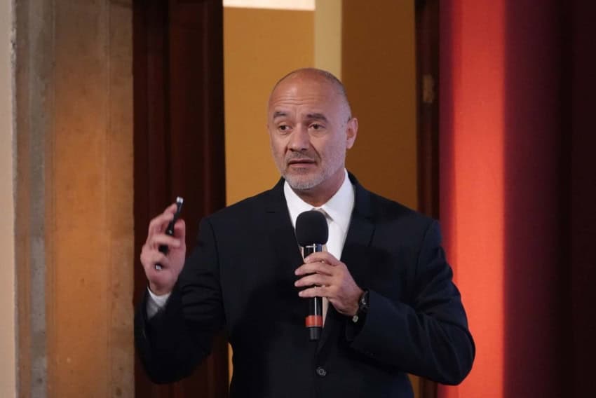 Mexico's Federal Infrastructure, Communications and Transport Minister Jesús Antonio Esteva Medina talking to an audience off camera while holding a small device in his right hand and a microphone in the other.