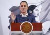 President Claudia Sheinbaum standing at the presidential podium in the National Palace during a press conference. She is wearing a blue blazer and a white turtleneck as she raises her right hand, palm outward, toward reporters as she speaks.