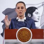 President Claudia Sheinbaum standing at the presidential podium in the National Palace during a press conference. She is wearing a blue blazer and a white turtleneck as she raises her right hand, palm outward, toward reporters as she speaks.