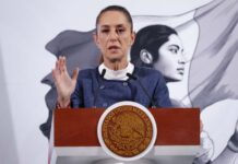 President Claudia Sheinbaum standing at the presidential podium in the National Palace during a press conference. She is wearing a blue blazer and a white turtleneck as she raises her right hand, palm outward, toward reporters as she speaks.