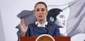 President Claudia Sheinbaum standing at the presidential podium in the National Palace during a press conference. She is wearing a blue blazer and a white turtleneck as she raises her right hand, palm outward, toward reporters as she speaks.