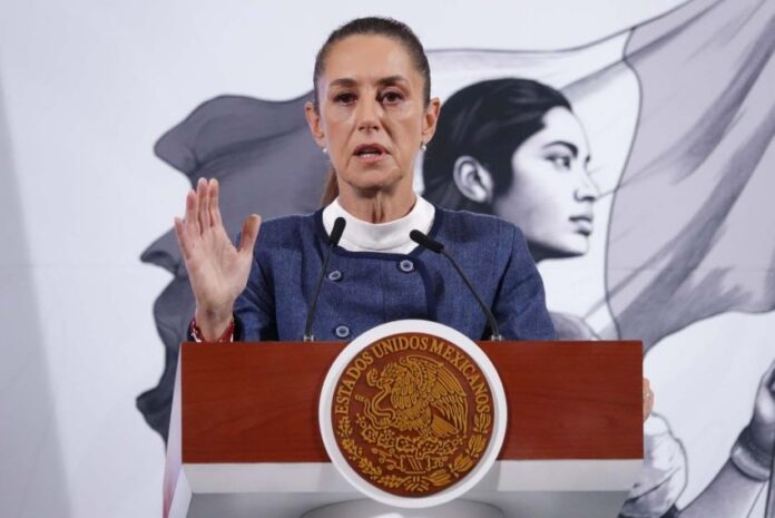 President Claudia Sheinbaum standing at the presidential podium in the National Palace during a press conference. She is wearing a blue blazer and a white turtleneck as she raises her right hand, palm outward, toward reporters as she speaks.