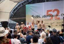 Band playing onstage at the International Cervantino Festival in 2024. The band of all men and one woman are dressed in traditional Mexican shirts, black pants and white campesino hats. One of the man is holding his hat up and out at the audience.