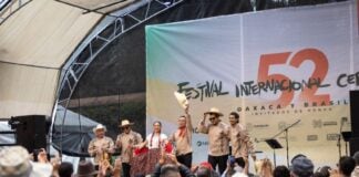 Band playing onstage at the International Cervantino Festival in 2024. The band of all men and one woman are dressed in traditional Mexican shirts, black pants and white campesino hats. One of the man is holding his hat up and out at the audience.