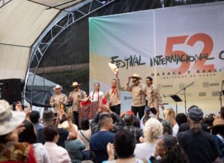 Band playing onstage at the International Cervantino Festival in 2024. The band of all men and one woman are dressed in traditional Mexican shirts, black pants and white campesino hats. One of the man is holding his hat up and out at the audience.