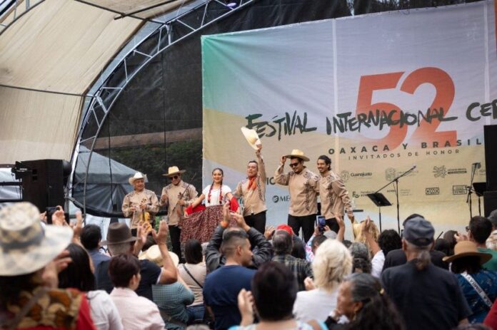 Band playing onstage at the International Cervantino Festival in 2024. The band of all men and one woman are dressed in traditional Mexican shirts, black pants and white campesino hats. One of the man is holding his hat up and out at the audience.