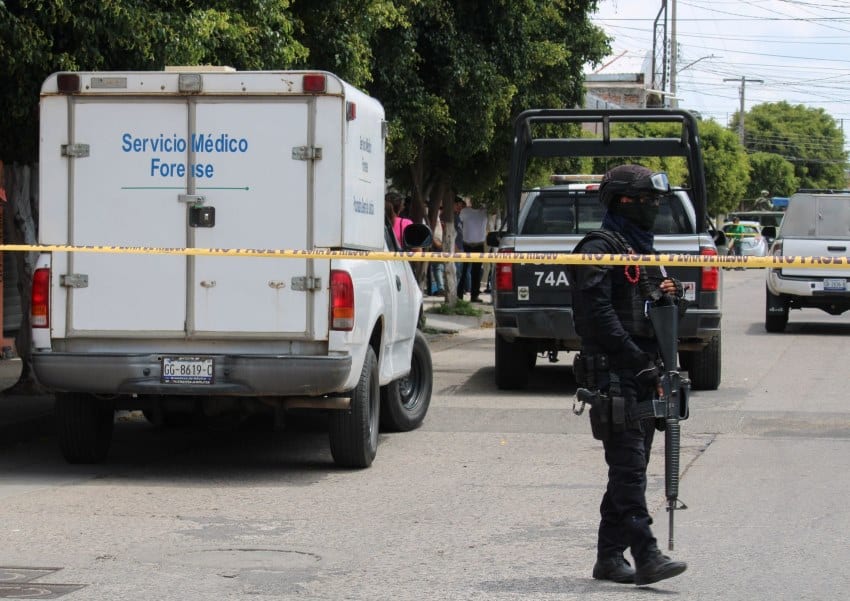 Police officer in black tactical gear and balaclava covering his face standing behind yellow police tape guarding a crime scene. In the background are police pickup trucks and a forensic services truck.