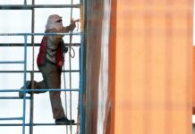 Man with his head and face covered with white cloths works on a building in Mexico City while standing on scaffolding next to the building.