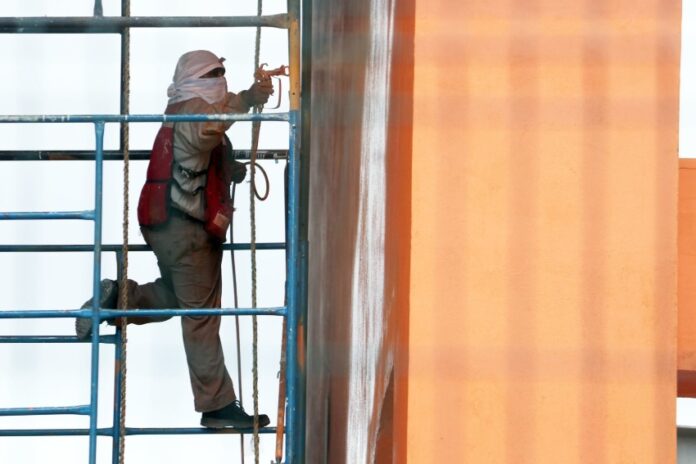 Man with his head and face covered with white cloths works on a building in Mexico City while standing on scaffolding next to the building.