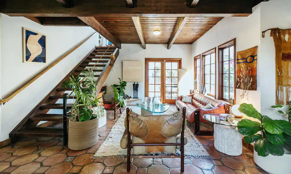 A well-lit sun room with French doors, an exposed-beam ceiling, plush leather furniture, large potted plants and artistic wall hangings