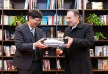 Two men opposite each other ,holding either side of a clear display box containing a toy-sized silver car. Behind them is a wall-sized bookcase with several shelves.