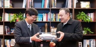 Two men opposite each other ,holding either side of a clear display box containing a toy-sized silver car. Behind them is a wall-sized bookcase with several shelves.