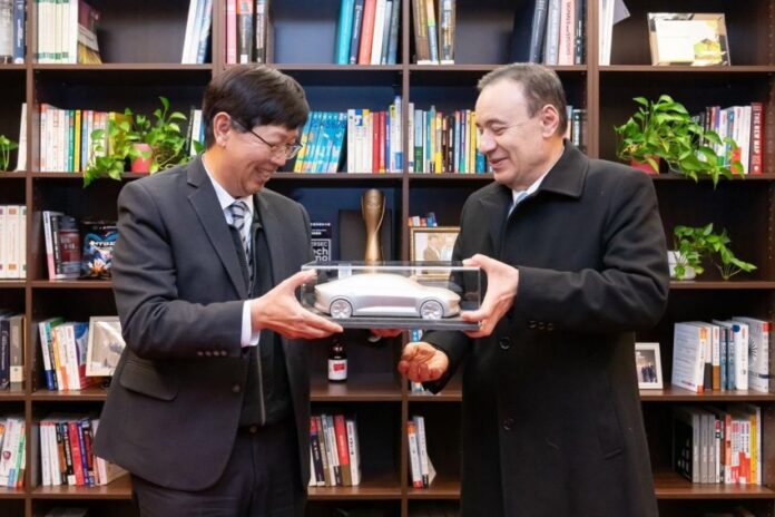 Two men opposite each other ,holding either side of a clear display box containing a toy-sized silver car. Behind them is a wall-sized bookcase with several shelves.