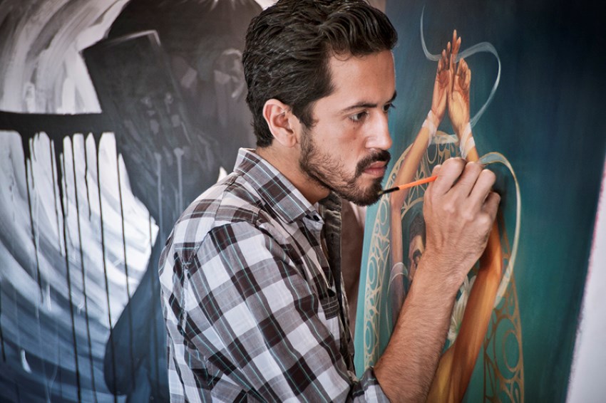 Mexican artist Fabian Chairez painting one of his works with a small brush. He is wearing a black and white plaid button down shirt and is staring intently at what he is doing. The painting takes up the entire background behind him.