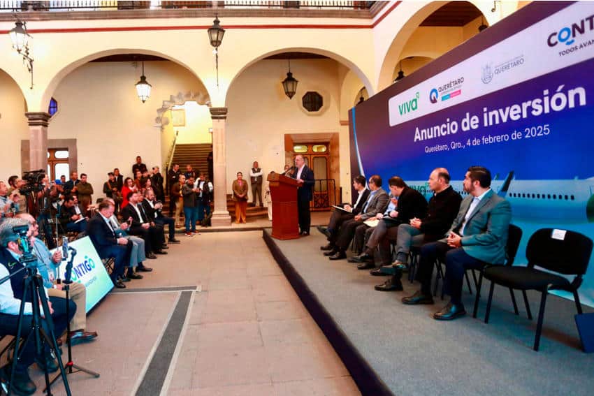 Reporters sitting as Gov. Mauricio Kuri gives a speech. On a dais are chairs with Queretaro and Viva Aerobus officials watching him speak.