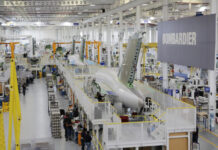 Bombardier aircraft parts manufacturing floor in Queretaro, Mexico, with a large sign that says "Bombardier" off to one side attached to support girders. A large fuselage is on a raised platform with workers at stations underneath.