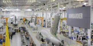 Bombardier aircraft parts manufacturing floor in Queretaro, Mexico, with a large sign that says "Bombardier" off to one side attached to support girders. A large fuselage is on a raised platform with workers at stations underneath.