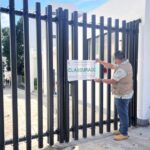 Man in a city government jacket putting a seal on a fenced gate to a hotel in Bacalar, Mexico. The seal says "Clausurado" (shut down)