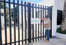 Man in a city government jacket putting a seal on a fenced gate to a hotel in Bacalar, Mexico. The seal says "Clausurado" (shut down)