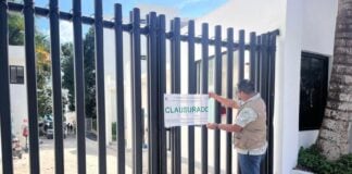 Man in a city government jacket putting a seal on a fenced gate to a hotel in Bacalar, Mexico. The seal says "Clausurado" (shut down)