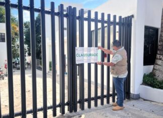 Man in a city government jacket putting a seal on a fenced gate to a hotel in Bacalar, Mexico. The seal says "Clausurado" (shut down)