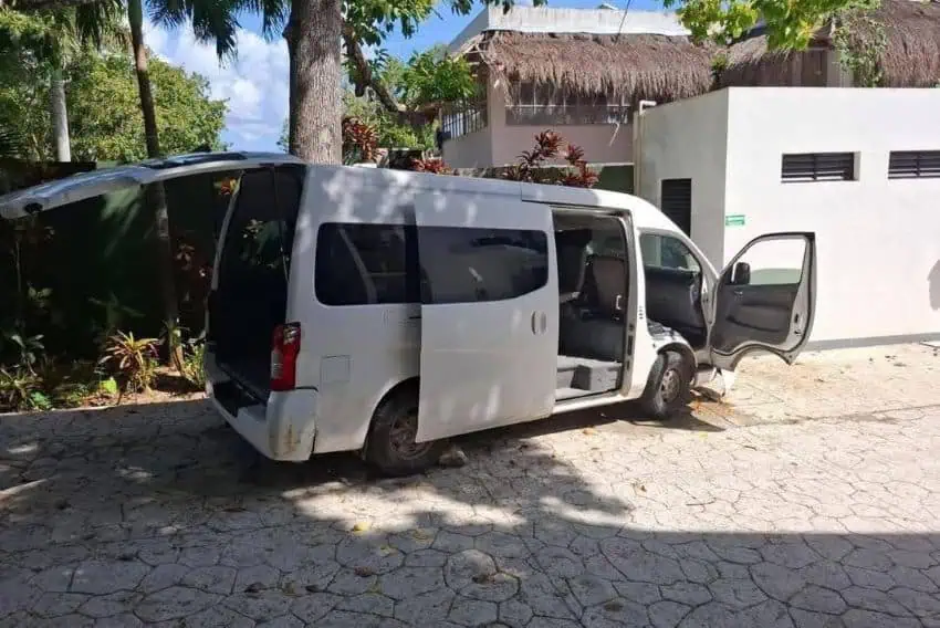 A white multiple passenger shuttle van parked on the pavement under some trees. All its doors are open.