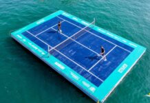 Aerial view of a regulation sized tennis court set up on wooden pallets, floating in the ocean and a man and a woman playing a match on the court.