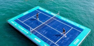 Aerial view of a regulation sized tennis court set up on wooden pallets, floating in the ocean and a man and a woman playing a match on the court.