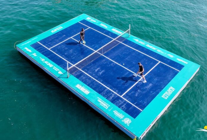 Aerial view of a regulation sized tennis court set up on wooden pallets, floating in the ocean and a man and a woman playing a match on the court.