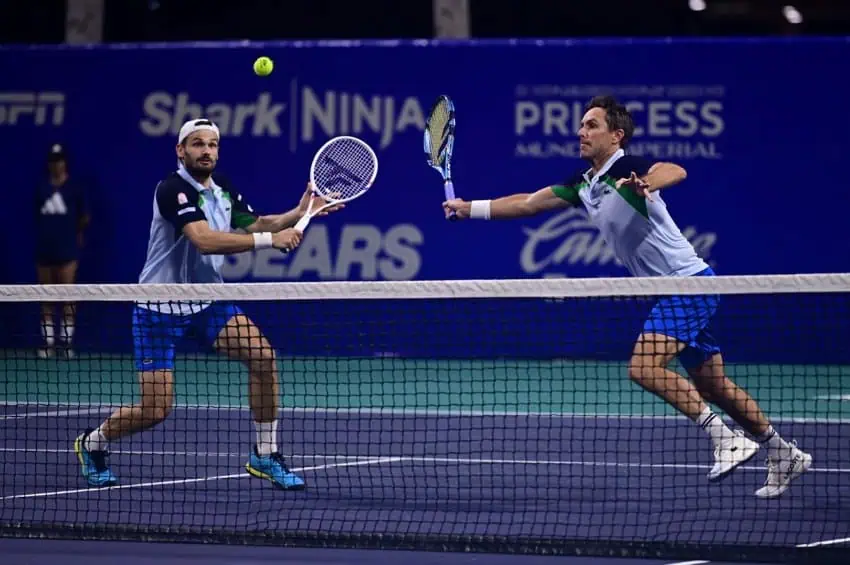 Two male tennis players on the same team in a doubles match in the 2025 Mexican Open in Acapulco. They are on a hard court, reaching with their racquets toward the same tennis ball flying in the air toward them.