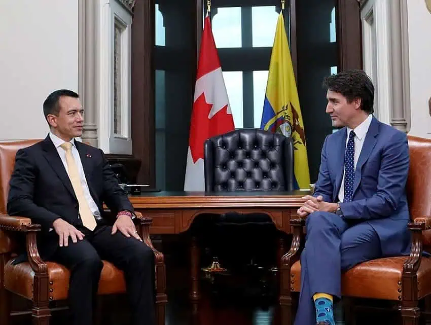 Ecuador's President Daniel Noboa seated across and to the side of Canada's Prime Minister Justin Trudeau. Each are in an antique style chair in front of a desk and chair, with the full-sized flags of Canada and Ecuador standing behind the chair. The two men are looking at each other. Trudeau haas his hands folded together in his lap while Noboa's hands are open, with one on each thigh, palms down. Both are smiling.