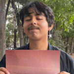 A young man with thick, ear-length hair and wearing an earring with a metal star stares at the camera with his eyes closed and holding a piece of cardboard on a sidewalk in Mexico City.