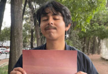 A young man with thick, ear-length hair and wearing an earring with a metal star stares at the camera with his eyes closed and holding a piece of cardboard on a sidewalk in Mexico City.