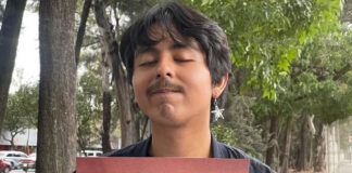 A young man with thick, ear-length hair and wearing an earring with a metal star stares at the camera with his eyes closed and holding a piece of cardboard on a sidewalk in Mexico City.