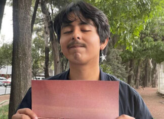 A young man with thick, ear-length hair and wearing an earring with a metal star stares at the camera with his eyes closed and holding a piece of cardboard on a sidewalk in Mexico City.