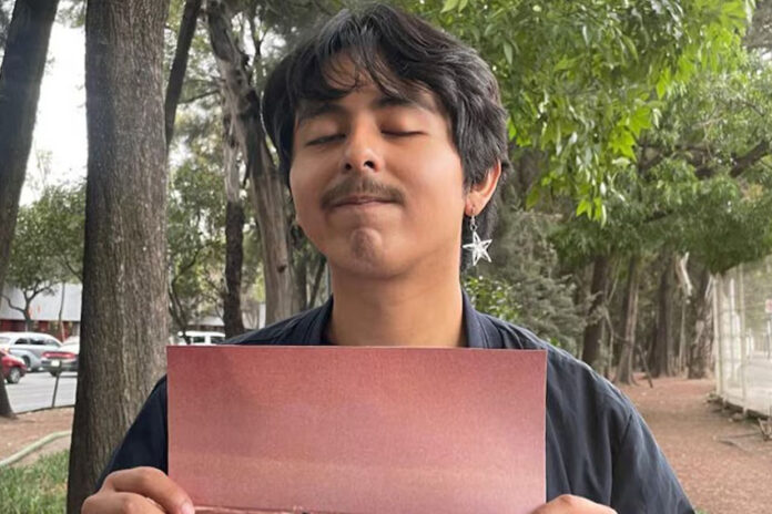 A young man with thick, ear-length hair and wearing an earring with a metal star stares at the camera with his eyes closed and holding a piece of cardboard on a sidewalk in Mexico City.