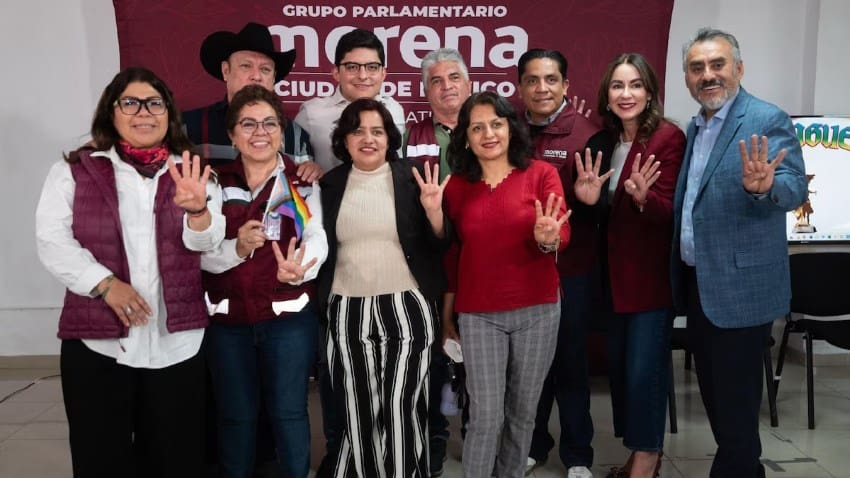 A group of Mexico City legislators and Morena Party functionaries posing in front of a banner for the Mexico City Morena Party. They are smiling and all holding up a hand with four fingers up.