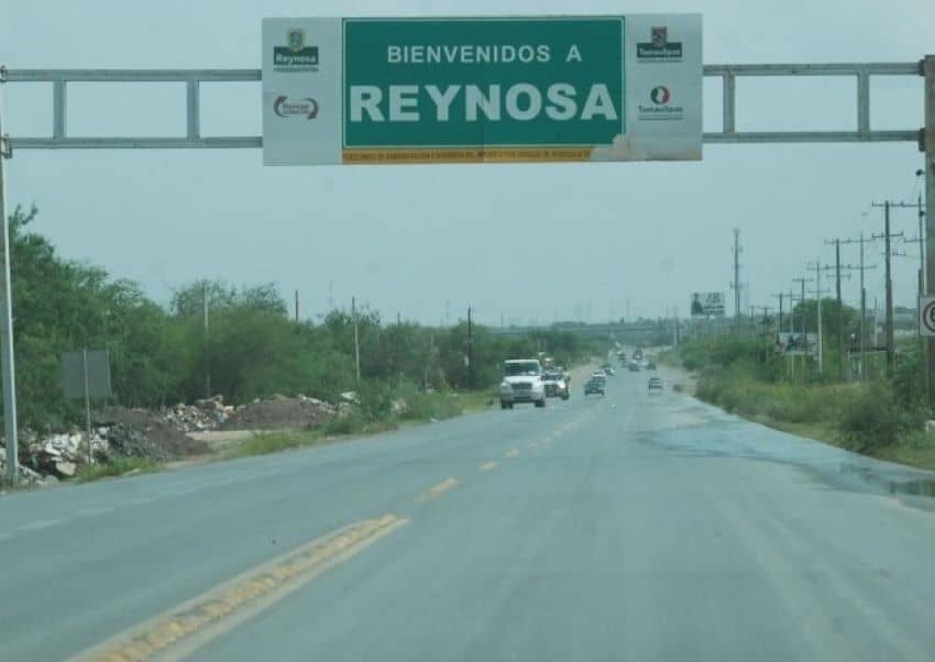 Image of a sparsely populated highway with a tractor trailer and other cars in the distance. In the foreground is a government highway sign proclaiming "Welcome to Reynosa" in Spanish.