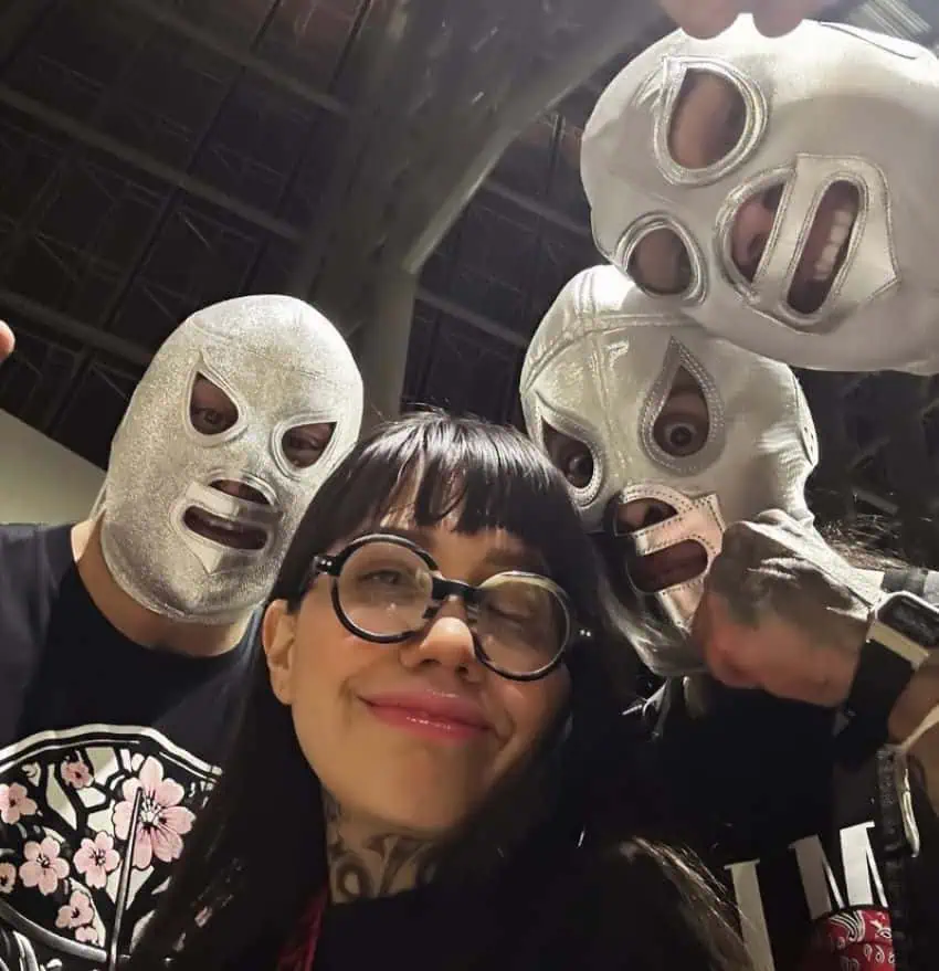 Author Dahlia de la Cerda posing with lucha libre legends El Hijo Santo, Santo Jr. and her boyfriend wearing a lucha libre mask in the style of the late fighter El Santo. All except de la Cerda are wearing the same style of silver lucha libre mask as they stare down into the camera.