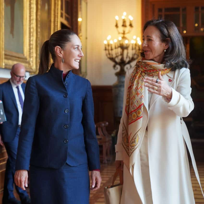 Mexico's President Claudia Sheinbaum walking next to Ana Botin, CEO of Grupo Santander inside Mexico's National Palace. Sheinbaum is wearing a matching royal blue skirt blazer set and Botin is wearing a long white coat and a large colorful scarf around her neck and draped over her right coat lapel. She is gesticulating at Sheinbaum, who looks back at her with her arms down at her sides and smiling.