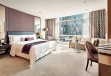 luxury hotel room in Mexico City with king sized bed in white with multiple pillows, a desk, sofa and coffee table and a view of a skyscraper across the street outside.