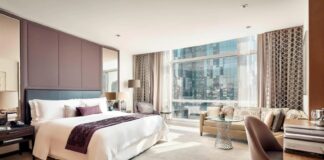 luxury hotel room in Mexico City with king sized bed in white with multiple pillows, a desk, sofa and coffee table and a view of a skyscraper across the street outside.