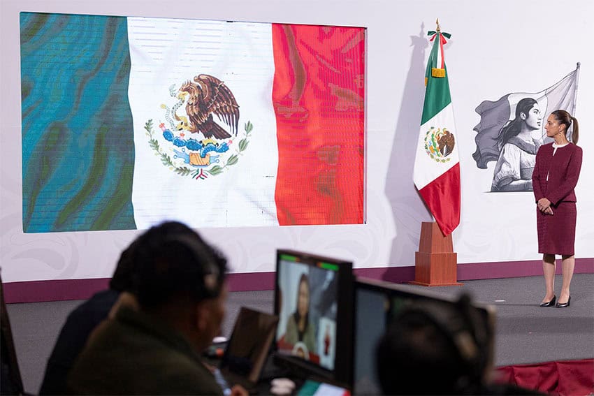 President Sheinbaum stands next to a large Mexican flag at her morning press conference, where she discussed Trump's tariff threats and the USMCA free trade deal