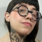 Dahlia de la Cerda in a publicity photo. The photo is a closeup of her face. She wears thick round black glasses and has long black hair and bangs. Some of her tattoos on her neck are visible.