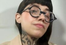 Dahlia de la Cerda in a publicity photo. The photo is a closeup of her face. She wears thick round black glasses and has long black hair and bangs. Some of her tattoos on her neck are visible.